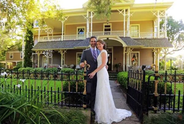 A bride and groom smile and embrace in front of a black iron gate, with a large, light-colored Victorian-style house and green garden in the background.
