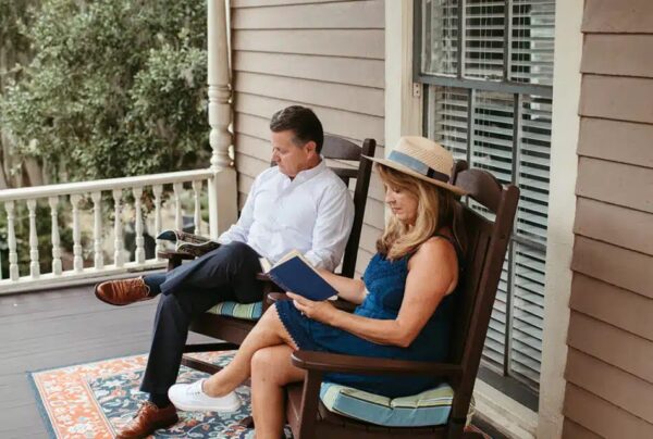 A man and woman sit in rocking chairs on a porch, reading books. The woman wears a blue dress and straw hat, and the man wears a white shirt and brown shoes. Theres a colorful rug on the wooden floor.