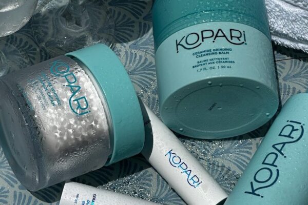 Close-up of several Kopari skincare products with light blue lids and labels, resting on a patterned surface. The containers are covered in water droplets, suggesting a fresh or spa-like environment.