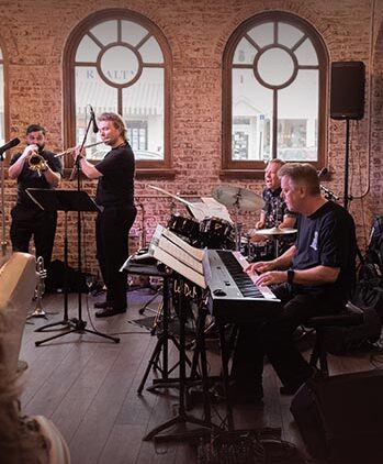 A jazz band performs indoors; three musicians stand playing brass instruments, while two others sit playing drums and keyboard. The room has brick walls, arched windows, and wooden floors.