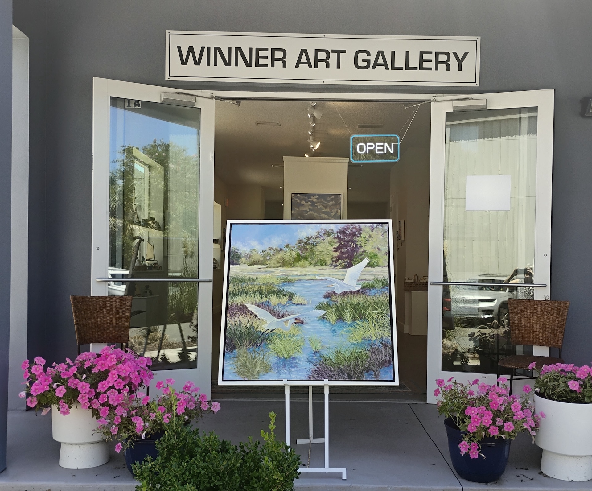 An art gallery entrance with open doors, pink flowers in pots, and a painting of white birds over a stream displayed on an easel. A glowing OPEN sign hangs above the door.